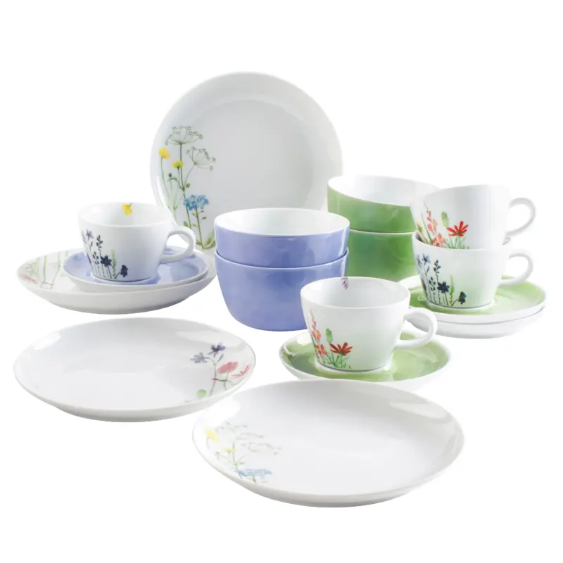 Eine helle, floral dekorierte Geschirrkollektion, bestehend aus mehreren Tellern, Untertassen und Tassen in verschiedenen Farben, darunter Blau und Grün. Die Motive zeigen zarte Blumenmuster auf weißem Porzellan. Die Tassen sind teilweise gestapelt, während die größeren Teller flach liegen. Diese Anordnung vermittelt einen fröhlichen, einladenden Eindruck.
